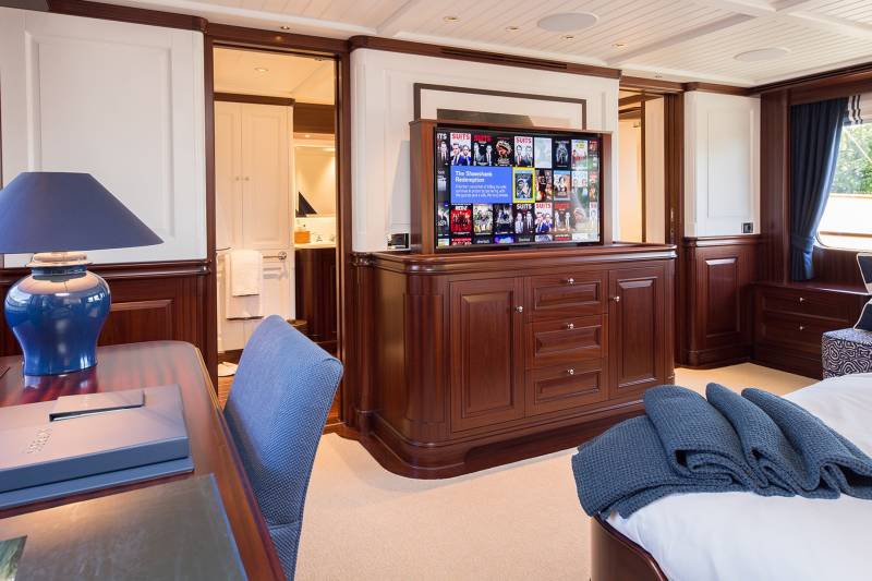 Luxurious yacht cabin features a large television displaying movie options, a comfortable desk with a lamp, and a plush bed with folded blankets.