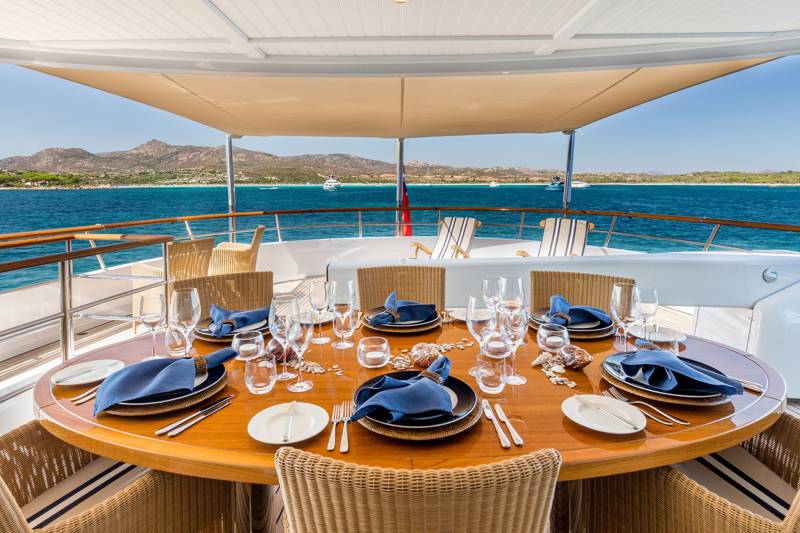 A beautifully set dining table on a yacht deck overlooks a stunning Mediterranean coastline.