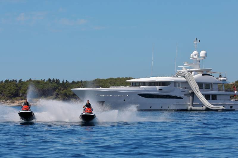 Two jet skiers create a splash as they ride past a luxurious yacht anchored near a tree-lined coast.