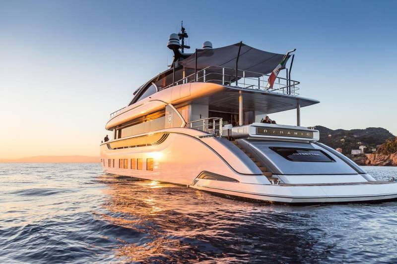 A luxurious yacht glides across the calm sea at sunset, reflecting the golden light and the distant Italian coastline.