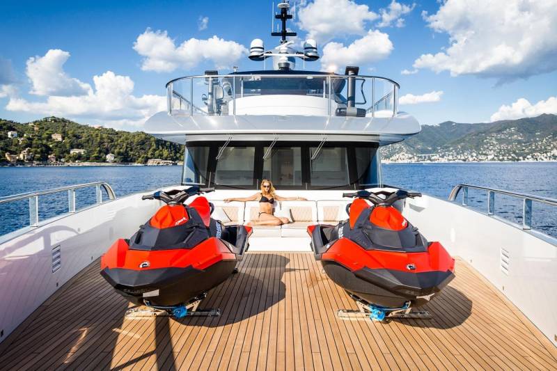 A woman relaxes on a luxury yacht's deck between two vibrant red jet skis, with a picturesque coastal town in the background.