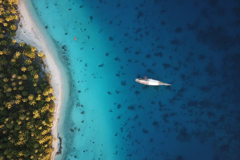 An aerial view captures a sleek yacht sailing through crystal-clear turquoise waters beside a lush palm-fringed island.