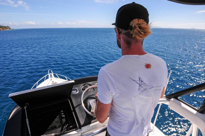 A man with a blonde man bun steers a boat through sparkling blue ocean waters under a clear sky.