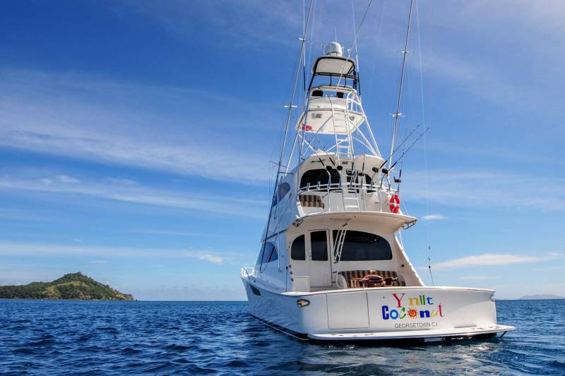 A luxurious yacht named "Ynlt Coconut" cruises through the sparkling blue waters under a bright, cloudy sky.