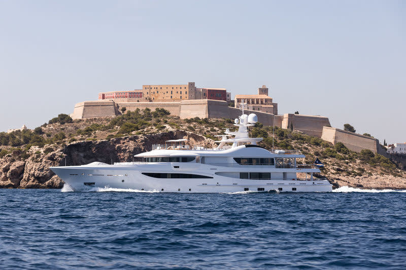 A sleek white superyacht glides through the azure waters with a historic fortress perched on the cliffside above.