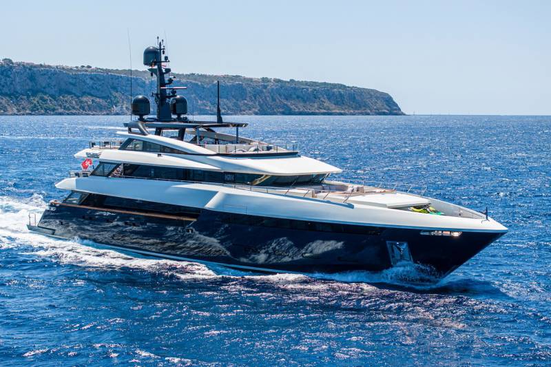 A sleek, modern yacht cruises through the sparkling blue waters off a rugged, sun-drenched coastline.