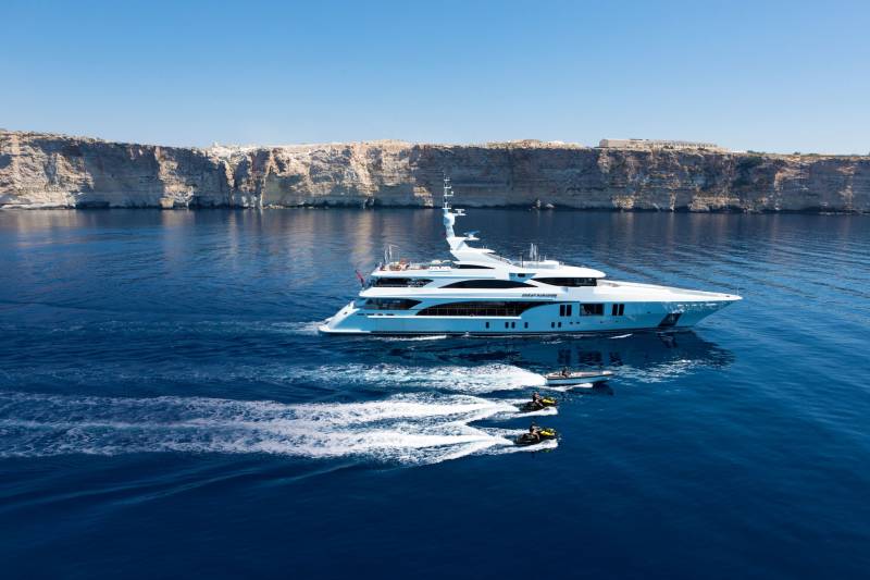 A luxurious yacht cruises through the azure waters near dramatic cliffs, trailed by jet skis and a tender.