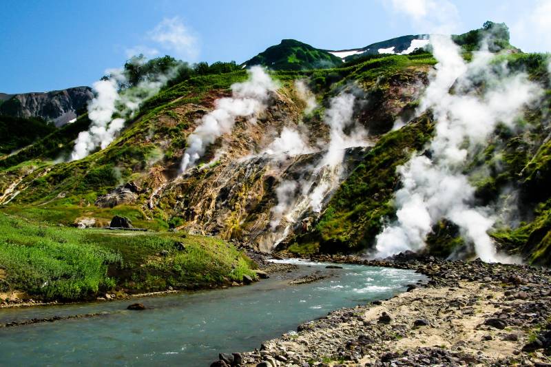 Steaming hot springs cascade down a vibrant green mountainside into a clear blue river.
