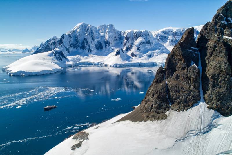 A research vessel navigates the icy waters of Antarctica, dwarfed by majestic snow-capped mountains.