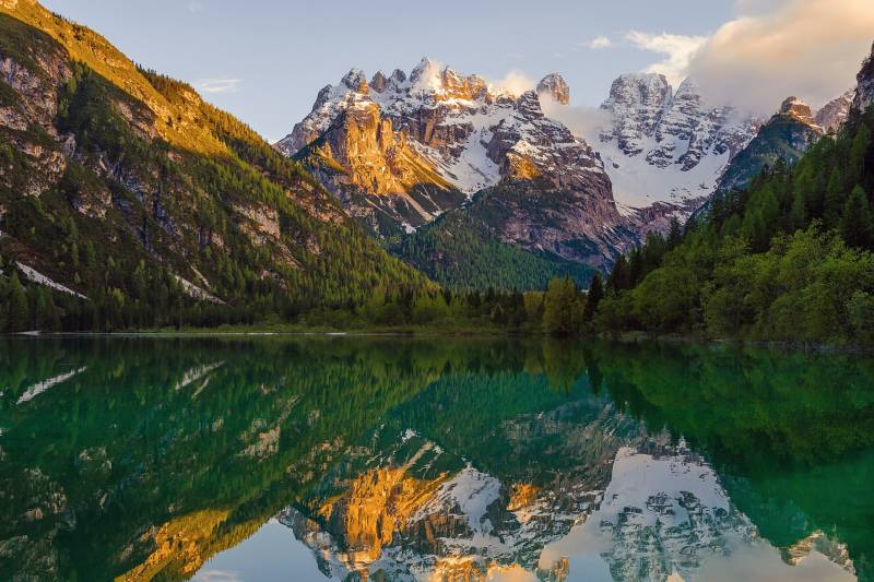 Golden sunlight illuminates snow-capped peaks reflected in the emerald waters of a serene mountain lake.