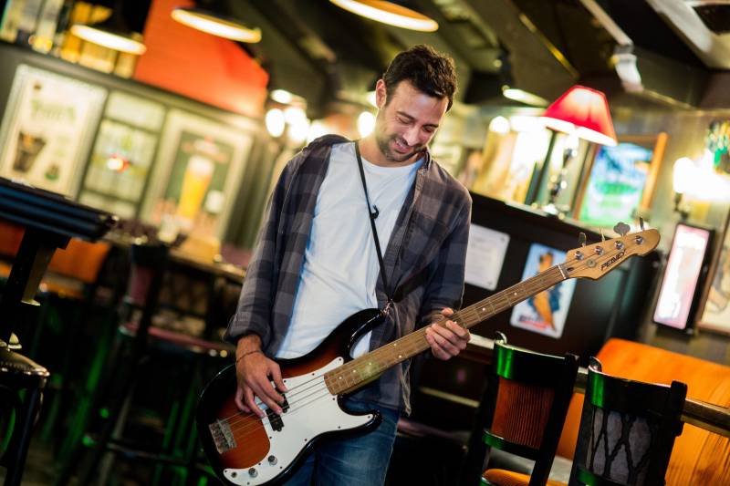 A man plays a bass guitar in a dimly lit bar, surrounded by vintage decor and warm lighting.