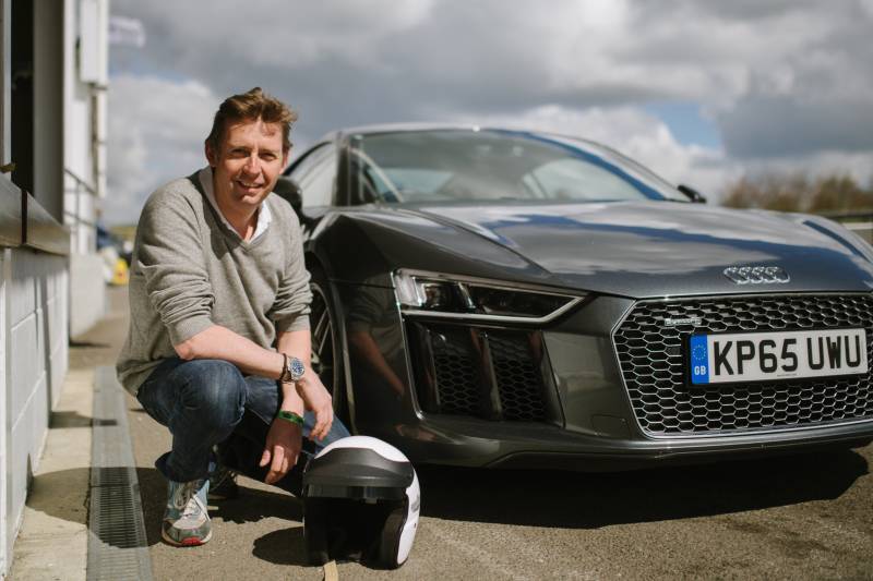A man in a grey sweater and jeans crouches beside a sleek grey Audi R8 on a sunny day.