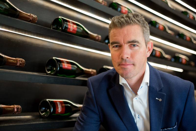 A man in a suit stands before a wall of champagne bottles, illuminated by soft, horizontal lights.