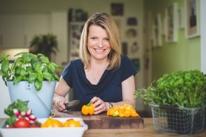 A smiling woman prepares fresh bell peppers and vegetables in a bright, homey kitchen.
