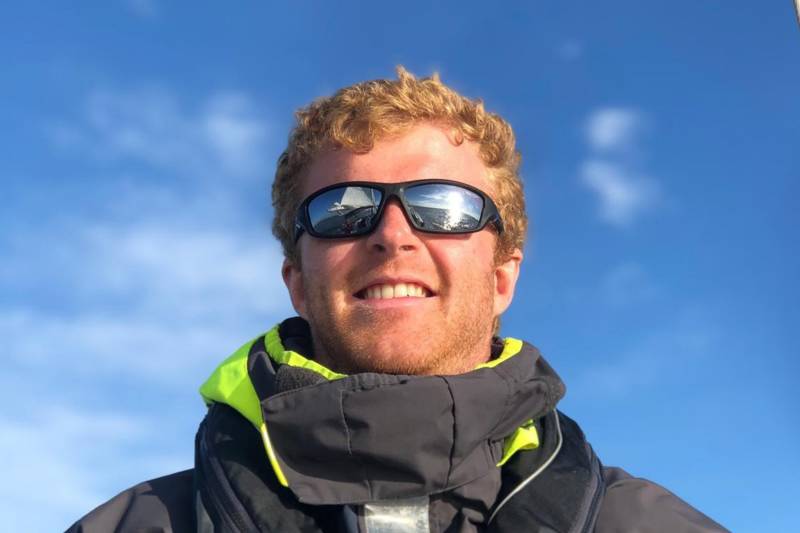 A smiling man with red hair and sunglasses reflects a sailboat on the water under a bright blue sky.