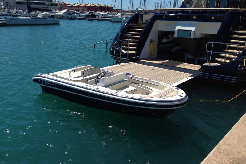 A small tender boat named "Mytrust Fund" floats serenely beside a large yacht docked in a vibrant blue harbor.