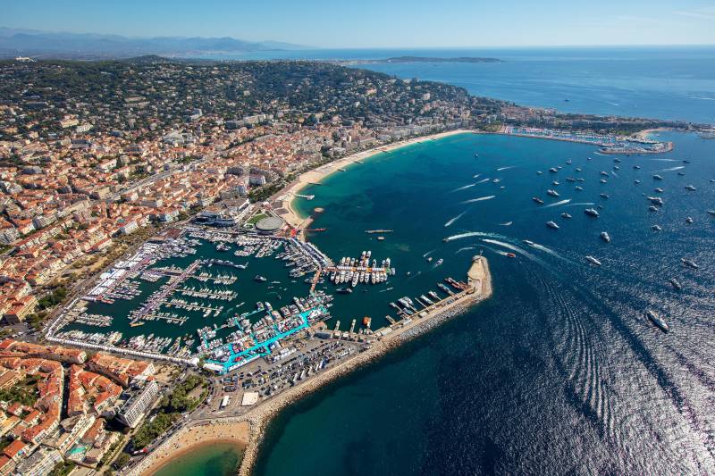 An aerial view captures the bustling marina and vibrant coastline of Cannes, France, dotted with numerous yachts under a clear blue sky.