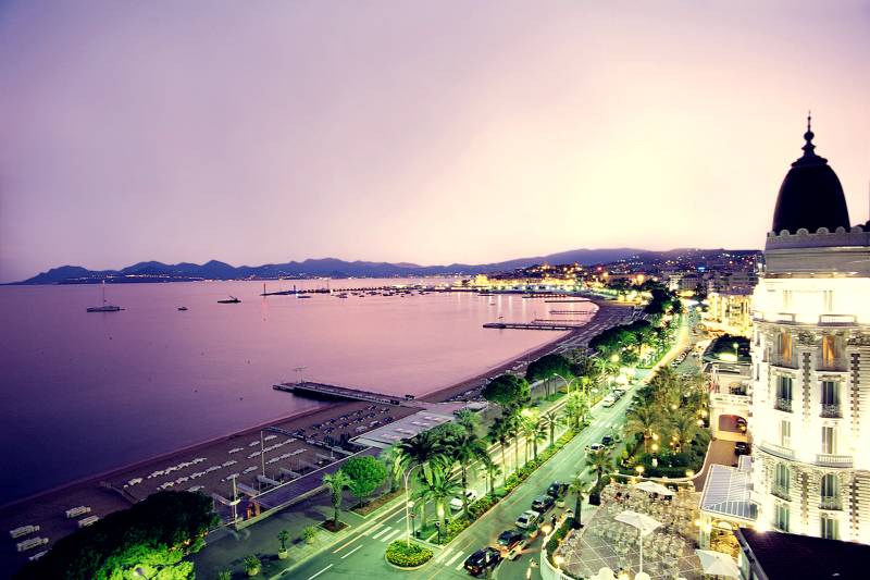 A coastal city glows with lights as dusk settles over the serene bay and palm-lined promenade.