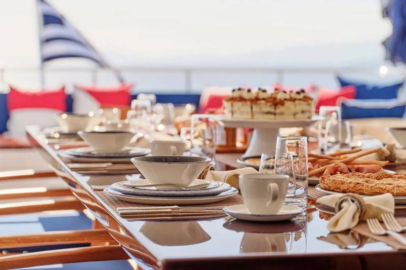 A beautifully set table on a yacht deck awaits a delicious meal with a cake and savory pastries.