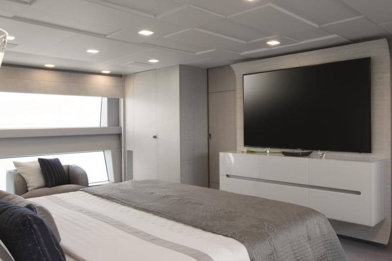 A modern and minimalist bedroom with a large television mounted on the wall, a white dresser, and a cozy-looking bed with gray bedding.