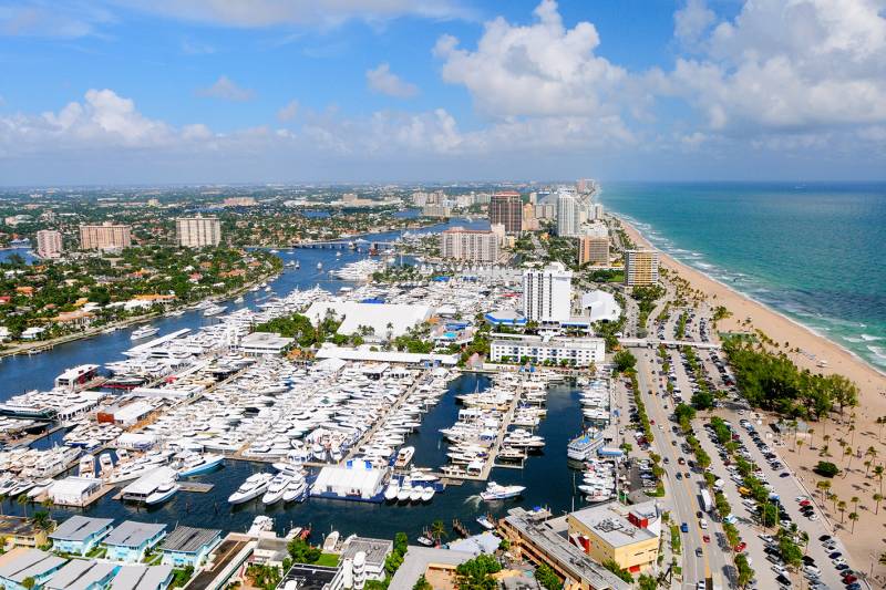 A bustling coastal city with a vibrant marina filled with boats, surrounded by high-rise buildings and lush greenery against a backdrop of a clear blue sky with fluffy white clouds.
