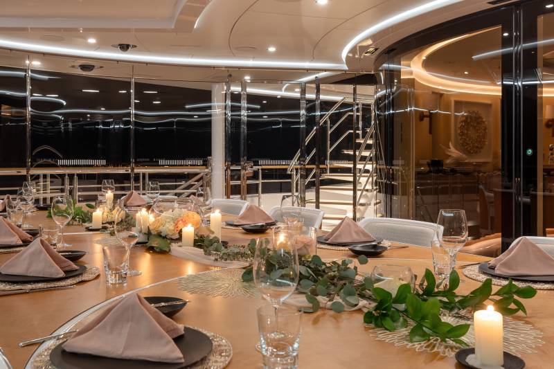 A beautifully set dining table on a yacht awaits guests with elegant place settings, floral arrangements, and flickering candles.