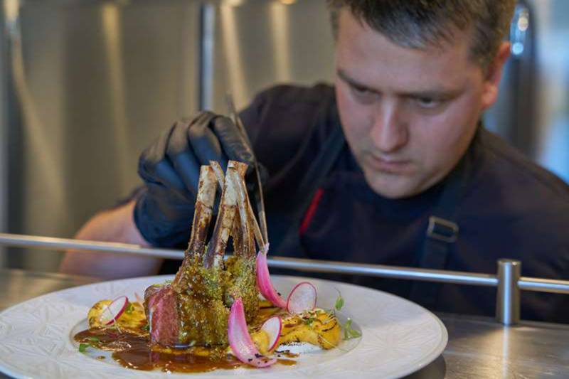 A chef meticulously garnishes a beautifully plated rack of lamb with thinly sliced radishes and herbs.