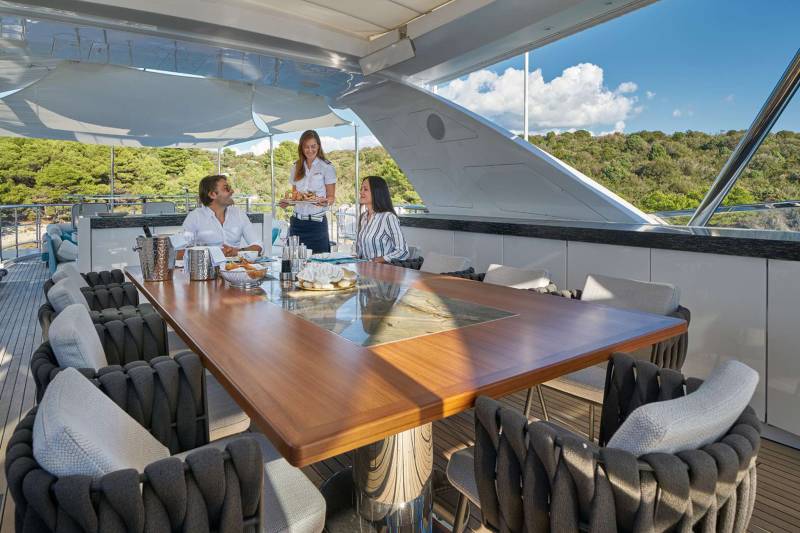 A server presents a platter of seafood to a couple dining al fresco on a luxurious yacht deck overlooking a verdant coastline.