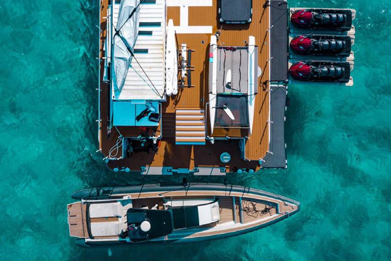 Three jet skis are docked alongside a large yacht in the clear turquoise waters of a tropical paradise.