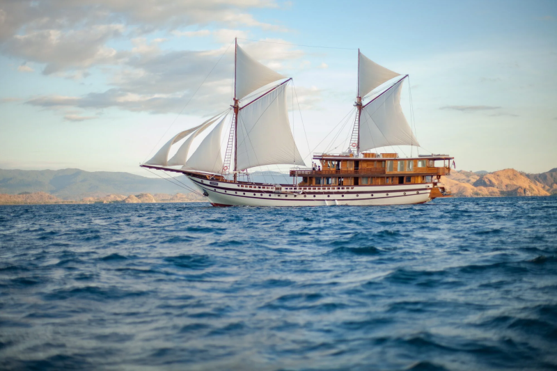 A majestic schooner glides across the azure sea, its white sails billowing against a backdrop of rugged, sun-kissed hills.