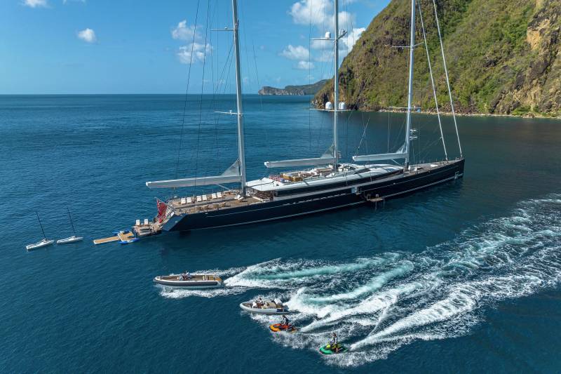A majestic three-masted schooner anchors offshore as jet skis and dinghies create white wakes in the azure waters near a lush, verdant island.