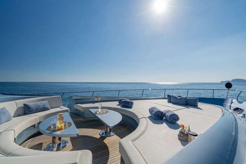 A luxurious yacht deck with comfortable seating arrangements, surrounded by the serene blue waters and a clear sky with a bright sun.