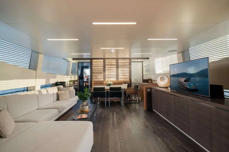 A modern and spacious living room with comfortable white sofas, a large television, and a wooden dining table with chairs, all set against a backdrop of large windows and natural lighting.