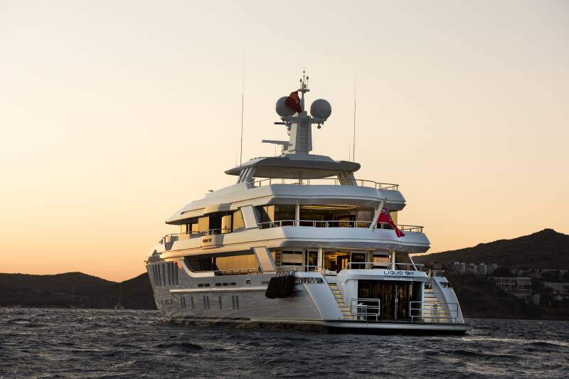 A luxurious yacht named Liquid Sky sails on calm waters under a warm, dusky sky near a coastal town.