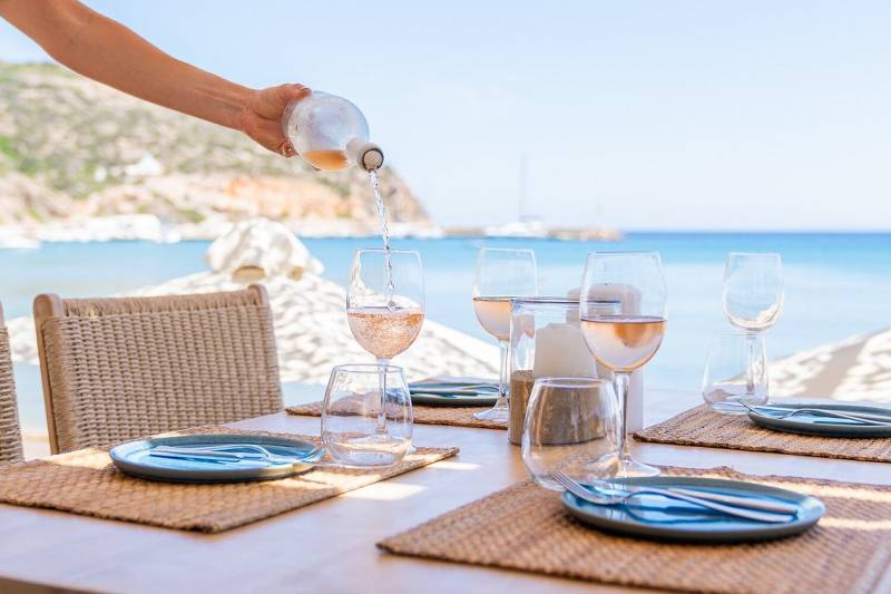 A hand pours rosé wine into a glass at a table set for dining with a stunning ocean view.
