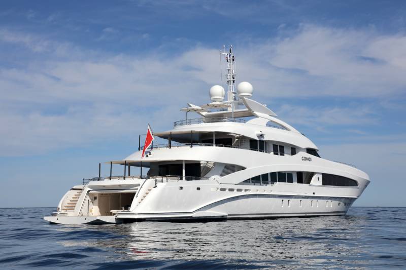 A luxurious white yacht named COMO glides serenely across the calm blue ocean under a bright, cloudy sky.