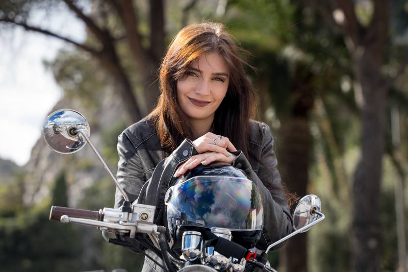 A stylish woman in a leather jacket rests her hands on a motorcycle helmet, her gaze meeting the camera with a confident smile.