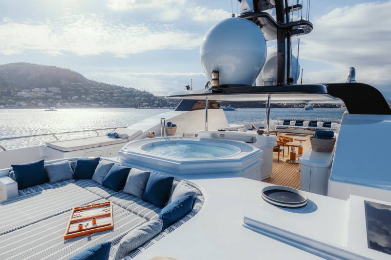 A luxurious yacht deck features a bubbling hot tub and comfortable seating overlooking a sun-drenched coastline.