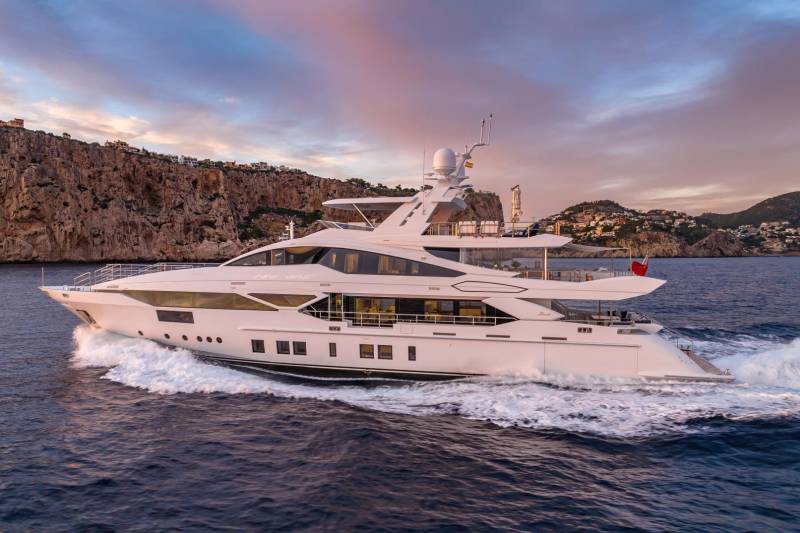 A luxurious white yacht cruises through the deep blue sea against a backdrop of rugged cliffs and a colorful sunset sky.