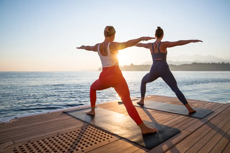 Two women practice yoga in warrior pose on a wooden deck overlooking a calm sea at sunrise.