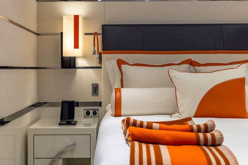 A modern bedroom features a plush bed with orange and cream accents, complemented by a sleek bedside table and ambient lighting.