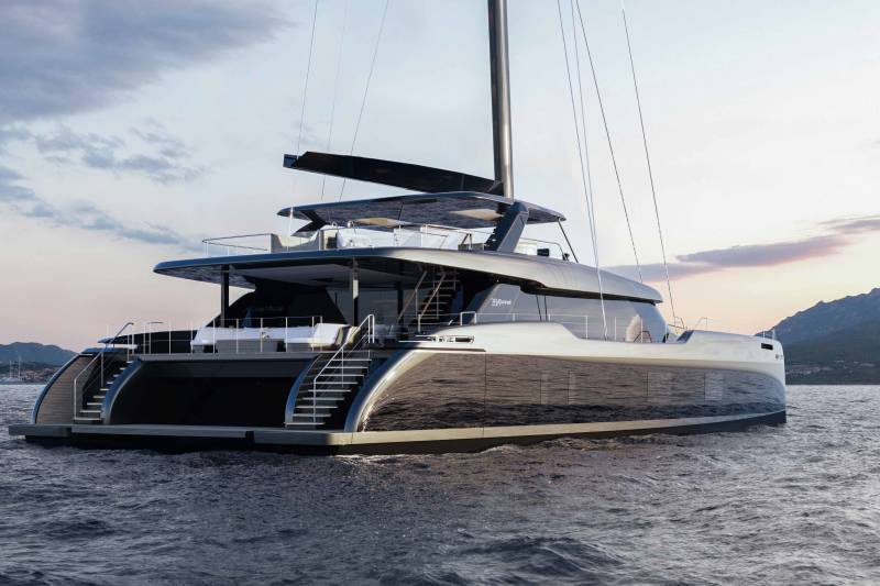 A sleek, modern catamaran yacht glides through the calm, dark waters with a mountainous coastline in the background.