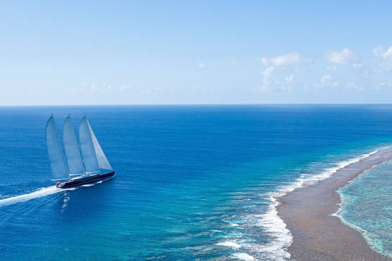 A majestic three-masted sailing yacht glides through the vibrant turquoise waters bordering a coral reef.