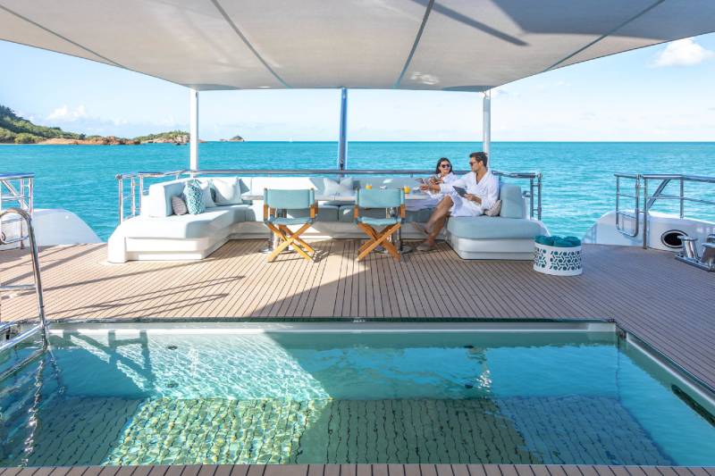 A couple relaxes on a luxurious yacht deck, enjoying drinks by a tranquil pool with a stunning ocean backdrop.