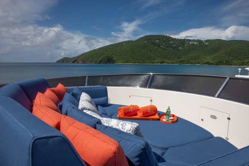 Luxurious blue and orange cushions await relaxation on a yacht deck overlooking a verdant, sun-drenched island.