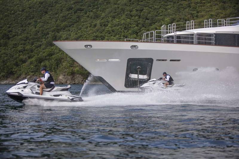 Two jet skiers race through the water alongside a large yacht against a lush green backdrop.