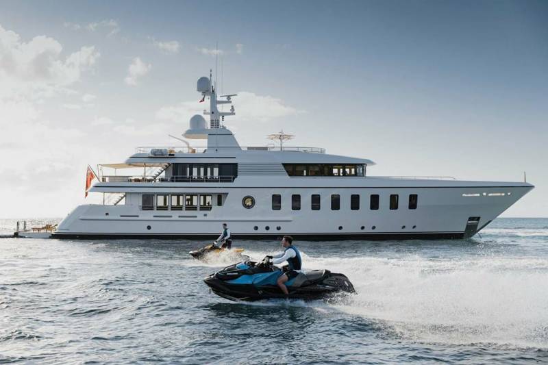 Two jet skiers race across the water in front of a large white yacht under a cloudy sky.