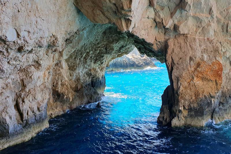 Sunlight illuminates the vibrant turquoise waters of a sea cave, framed by rugged, textured rock formations.