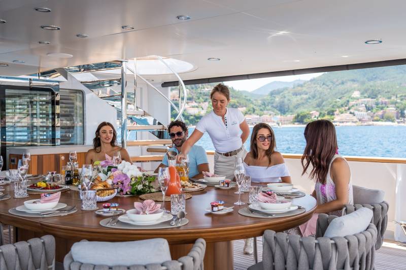 A group enjoys a luxurious al fresco meal on a yacht, with a server pouring drinks against a backdrop of a picturesque coastal town.
