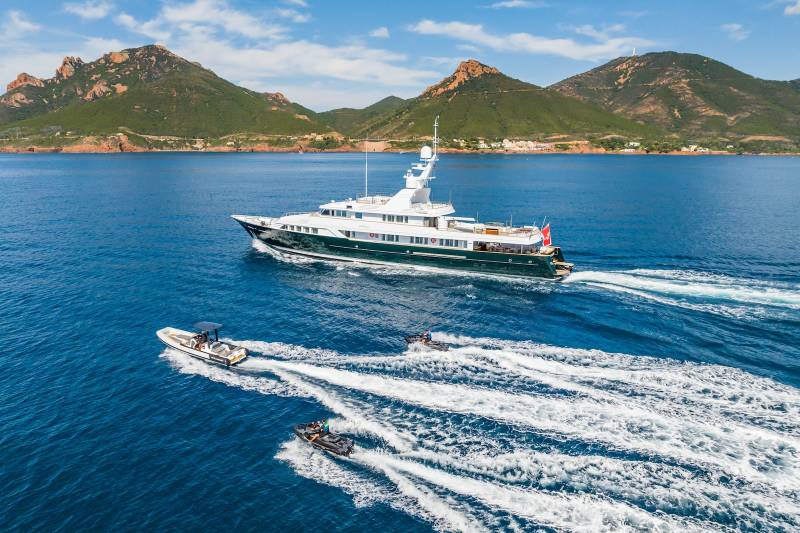 A luxurious yacht cruises through the azure waters, trailed by jet skis and a tender, with a dramatic mountainous coastline in the background.